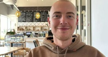 Portrait of a young man with a shaved head, smiling, wearing a tan hoodie.