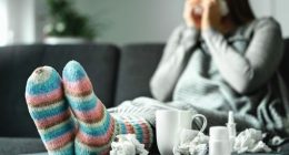 Sick woman with flu, cold, fever and cough sitting on couch at home. Ill person blowing nose and sneezing with tissue and handkerchief. Woolen socks and medicine. Infection in winter. Resting on sofa.