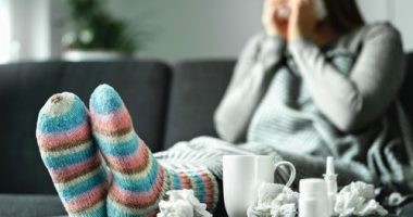 Sick woman with flu, cold, fever and cough sitting on couch at home. Ill person blowing nose and sneezing with tissue and handkerchief. Woolen socks and medicine. Infection in winter. Resting on sofa.