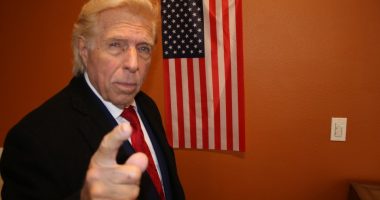 A Donald Trump doppelgänger points at the viewer in front of an American flag.