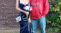 A father and daughter posing for a photo.  The daughter is wearing a dark blue dress and carrying a bouquet.