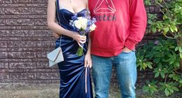 A father and daughter posing for a photo.  The daughter is wearing a dark blue dress and carrying a bouquet.