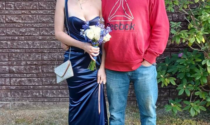 A father and daughter posing for a photo.  The daughter is wearing a dark blue dress and carrying a bouquet.