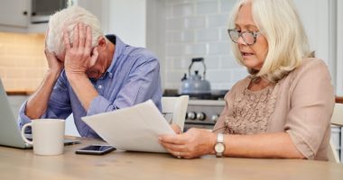Senior couple reviewing financial documents at their kitchen table, looking distressed.