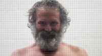 Shirtless man with long gray hair and beard behind glass.