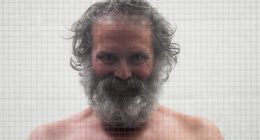 Shirtless man with long gray hair and beard behind glass.