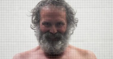 Shirtless man with long gray hair and beard behind glass.