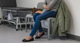 Full length wide view of a mid adult woman sitting in a doctors examination room waiting to be seen, she is holding her arm and looking up, feeling worried. She is wearing casual clothing at the hospital in Newcastle, England.