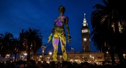 Illuminated sculpture of a woman projected onto a building at night, with spectators and palm trees in the foreground.