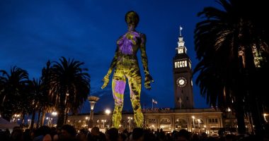 Illuminated sculpture of a woman projected onto a building at night, with spectators and palm trees in the foreground.