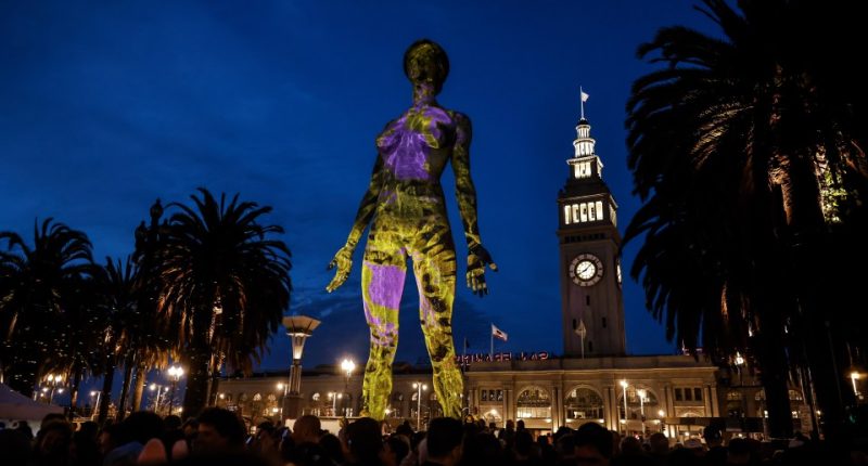 Illuminated sculpture of a woman projected onto a building at night, with spectators and palm trees in the foreground.