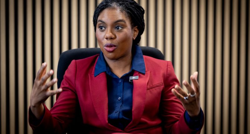 Photo of Kemi Badenoch in a red suit jacket, giving an interview.