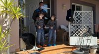 A masked man stands with two masked children on a porch, surrounded by clutter.