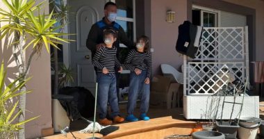 A masked man stands with two masked children on a porch, surrounded by clutter.