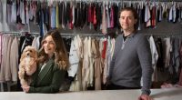 a woman holding a dog stands next to a man in front of clothes racks