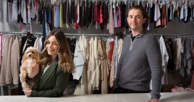 a woman holding a dog stands next to a man in front of clothes racks