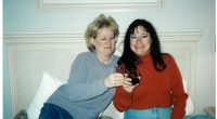 Two women toasting with drinks on a bed.