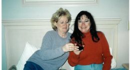 Two women toasting with drinks on a bed.