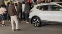 Screenshot of a crowd of people gathered around a bus and a white SUV.