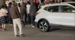 Screenshot of a crowd of people gathered around a bus and a white SUV.