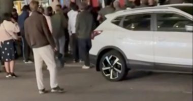 Screenshot of a crowd of people gathered around a bus and a white SUV.