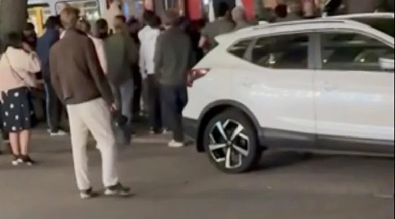 Screenshot of a crowd of people gathered around a bus and a white SUV.