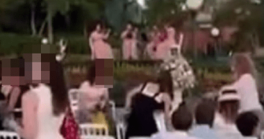People seated at white chairs outdoors watching a performance.