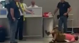 Person lying on the floor at an airport; several people standing nearby.
