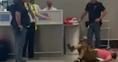 Person lying on the floor at an airport; several people standing nearby.