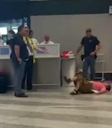 Person lying on the floor at an airport; several people standing nearby.