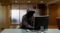A hooded figure sitting at a desk in an office, using a computer.