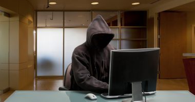 A hooded figure sitting at a desk in an office, using a computer.