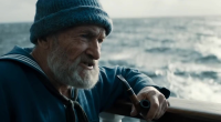 A medium shot frames an old sailor, his knitted blue sailor hat casting a shadow over his eyes, a thick grey beard obscuring his chin. He holds his pipe in one hand, gesturing with it towards the churning, grey sea beyond the ship's railing. "This ocean, it's a force, a wild, untamed might. And she commands your awe, with every breaking light."