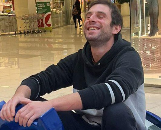 A smiling man sitting on a blue bumper car in a shopping mall.