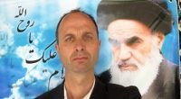 A man in a suit posing with a large photo of Ayatollah Khomeini in the background.