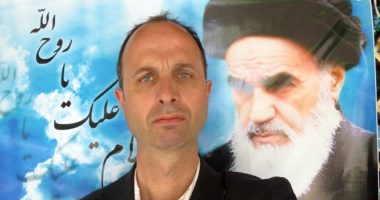 A man in a suit posing with a large photo of Ayatollah Khomeini in the background.