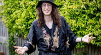 Man with long curly hair, glasses, and a black robe with gold embroidery, smiles and gestures with his hands.