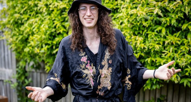Man with long curly hair, glasses, and a black robe with gold embroidery, smiles and gestures with his hands.