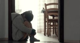 A sad young girl sits on the floor hugging her knees.