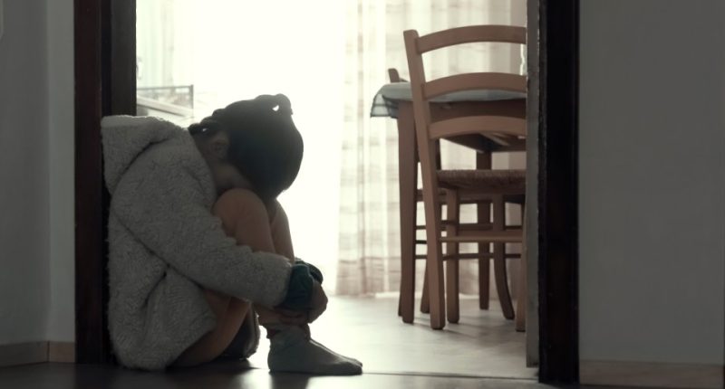 A sad young girl sits on the floor hugging her knees.