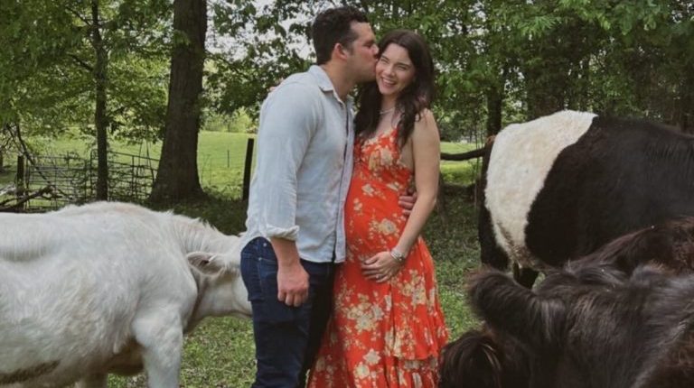 Pregnant woman in orange dress being kissed by a man, with cattle nearby.