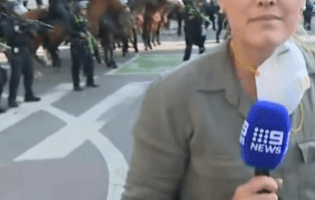 Reporter reporting live from a street with mounted police in the background.