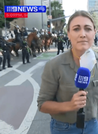 Reporter reporting live from a street with mounted police in the background.