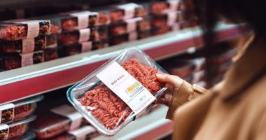 Above the shoulder view of woman holding a pack of beef mince in a supermarket. Standing in front of the meat product aisle and have no idea which product to choose from. The Conscious Carnivore - find meat that is locally-sourced, healthy, high quality butchering, and handled with respect. The environmentally conscious way to choosing food.