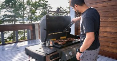 Man grilling meat and vegetables on a patio overlooking a lake.
