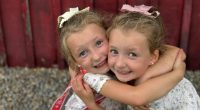 Twin girls hugging in front of a red wooden wall.