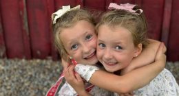 Twin girls hugging in front of a red wooden wall.