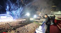 Kim Jong Un waves to a large crowd at a nighttime ceremony with fireworks.