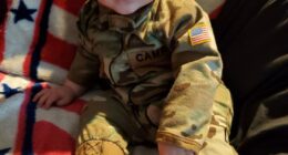 Baby in camouflage outfit on American flag blanket.
