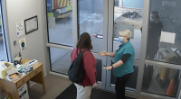Two people wearing masks greet each other at a doorway.